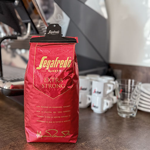 eine Packung Segafredo Extra Strong verpackt mit Aromaklammer vor einer Kaffeemaschine und einigen Tassen